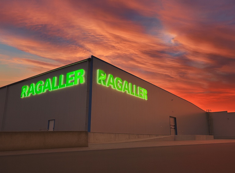 Ragaller-Logistikhalle in Hermsdorf mit leuchtendem grünen Schriftzug bei Sonnenuntergang – Symbol für modernes Qualitätsmanagement und nachhaltige Unternehmensführung.