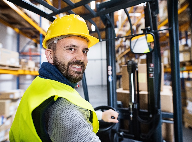 Lagerarbeiter mit Schutzhelm und Warnweste sitzt auf einem Gabelstapler und lächelt